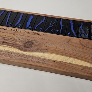 Puede incluir: Un tablero de juego de madera con las palabras "Finger Lakes Tile Game" grabadas en él. El tablero tiene 11 fichas numeradas con contornos azules, y un mapa de la región de los Finger Lakes está impreso en la parte inferior. Dos dados blancos están en el tablero.