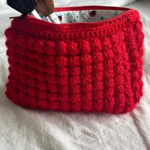 May include: A bright red crocheted pouch with a textured bobble stitch pattern and a zipper closure. The interior lining is white fabric featuring a pattern of small red ladybugs and teal dots. Two makeup brushes are tucked inside the pouch.