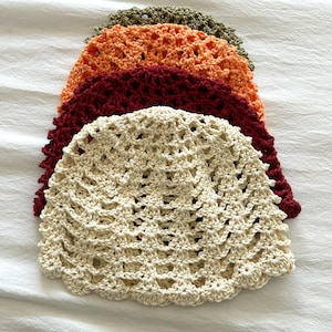 May include: A stack of four crocheted beanies in a shell stitch pattern. The hats are layered from top to bottom in olive green, bright orange, deep burgundy, and cream. Each beanie features a scalloped edge and a lightweight, open-weave design.
