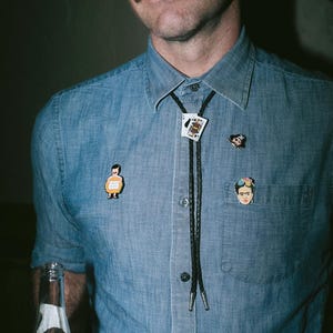 May include: A denim button-down shirt adorned with several pins and a bolo tie. The bolo tie features a playing card design. Additional pins include a cartoon figure, a Frida Kahlo portrait, and a small black and white pin.