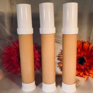 May include: Three tubes of beige-colored foundation with white caps. The tubes are arranged in a row on a surface with flowers in the background.