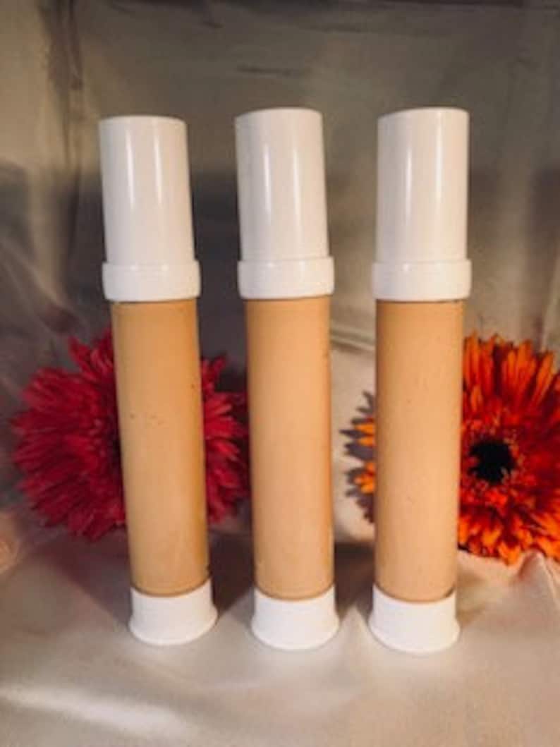 May include: Three tubes of beige-colored foundation with white caps. The tubes are arranged in a row on a surface with flowers in the background.