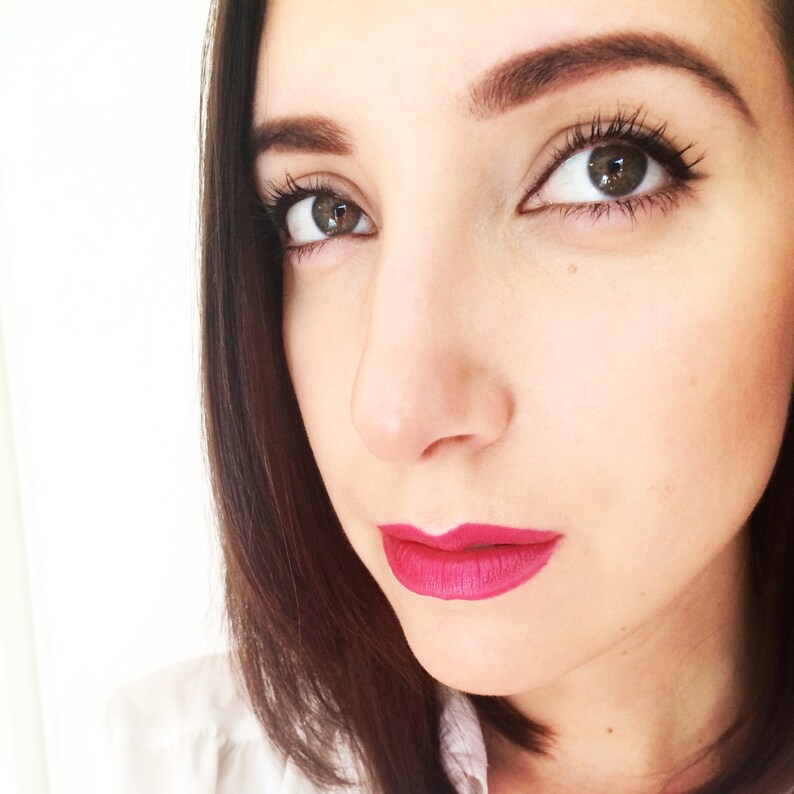 May include: A close-up of a woman's face with dark brown hair, defined eyebrows, and dark eyeliner. She is wearing bright pink lipstick.