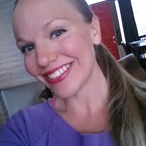 May include: A woman with long blonde hair is smiling and wearing a purple shirt.