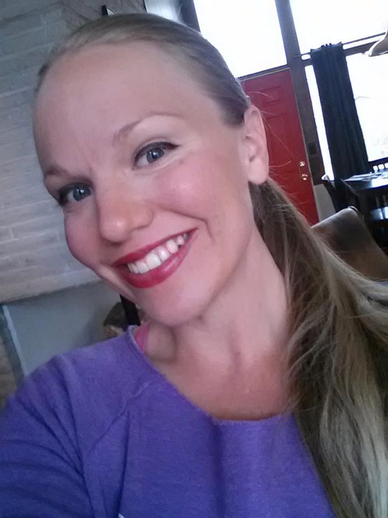 May include: A woman with long blonde hair is smiling and wearing a purple shirt.