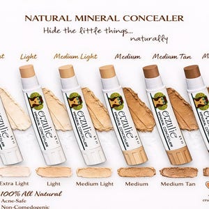 Könnte beinhalten: Präsentation von sechs Mineral-Concealer-Stiften in verschiedenen Farbtönen von Extra Light bis Medium Dark. Jeder weiße Stift trägt das "erzulie"-Logo und wird von einem Farbmuster der entsprechenden Farbe begleitet. Der Text "NATURAL MINERAL CONCEALER" steht oben.