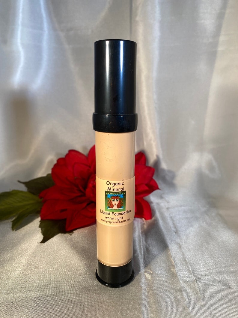 May include: A black tube of organic mineral liquid foundation in the shade warm light. The tube has a label with a cartoon woman and the text "Organic Mineral Liquid Foundation warm light www.progressinhealth.com".