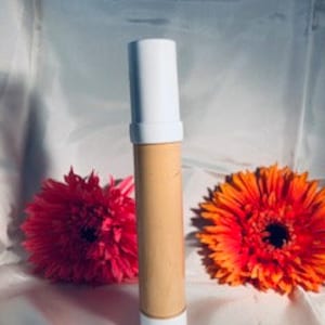 May include: A white tube with a beige liquid inside. The tube is standing upright on a surface with two pink and orange flowers behind it.