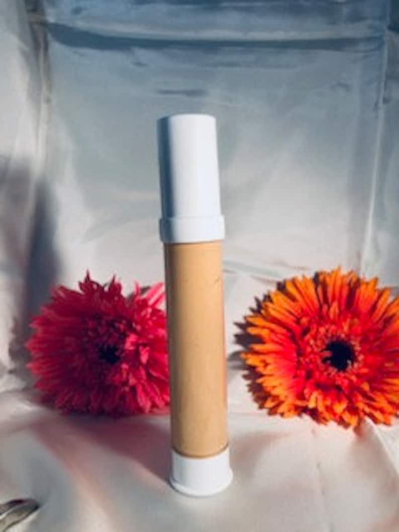 May include: A white tube with a beige liquid inside. The tube is standing upright on a surface with two pink and orange flowers behind it.