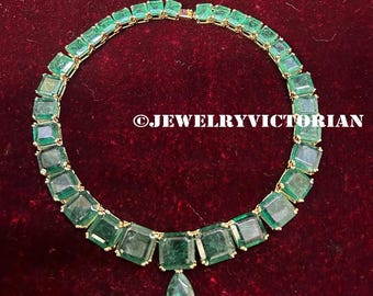Op de afbeelding: Een ketting met een reeks vierkant geslepen groene stenen in een gouden zetting, gerangschikt in een cirkelvormig patroon met een enkele peervormige groene hanger in het midden. De tekst JEWELRYVICTORIAN staat in het wit in het midden.