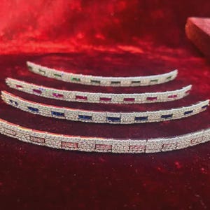 May include: Four curved, silver-toned bracelets with colorful rectangular gemstones. The gemstones are arranged in a repeating pattern of pink, blue, red, and green. The bracelets are displayed on a red velvet surface.
