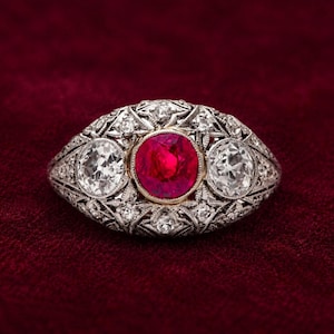 May include: A silver-toned ring featuring a central round-cut vibrant red ruby flanked by two round-cut clear diamonds. The metal band is intricately detailed with a filigree pattern and accented with numerous small, sparkling white stones.