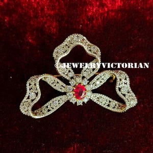 May include: A bow-shaped brooch featuring a central oval-cut red gemstone surrounded by a halo of small clear stones. The ribbon-like structure is encrusted with numerous clear stones and displays the text JEWELRYVICTORIAN across the center.