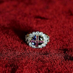 Puede incluir: Un anillo de oro con un diseño de cruz central, con piedras preciosas rojas y azules, rodeado por un halo de piedras blancas. El anillo está sobre un fondo rojo texturizado, creando un contraste llamativo.