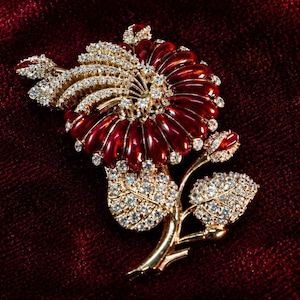 Può includere: Spilla floreale dorata con un fiore in smalto rosso e scintillanti cristalli trasparenti. La spilla presenta un design dettagliato di petali, foglie e uno stelo, su uno sfondo di velluto rosso scuro.