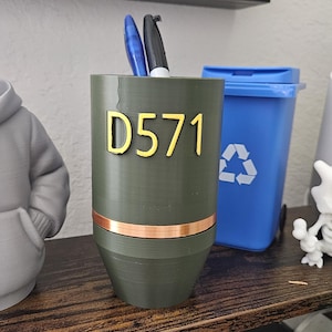 May include: An olive green pen holder with the text "D571" in gold. A copper-colored band encircles the middle. Two pens are visible inside. A blue recycling bin is in the background.