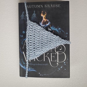 May include: A light blue crocheted bookmark in the shape of a triangle is placed on a book titled "The Wicked" by Autumn Krause. The book cover features a black background with a silhouette of a woman in a dress.