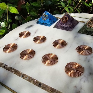 May include: A set of 12 copper wire spirals arranged in a grid pattern on a white marble surface. Four crystal pyramids with gold and silver swirls are arranged in a row above the spirals. The pyramids are made of blue, purple, green, and red crystals.