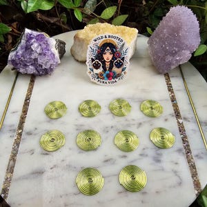 May include: A white marble tray with a gold trim and a sticker that says "Wild & Rooted, Pura Vida". The sticker features a woman with long dark hair and two black cats. There are 10 green metal coils on the tray.