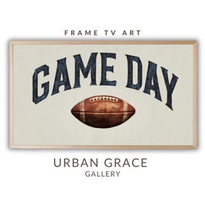 Puede incluir: Impresión en lienzo enmarcada con las palabras "GAME DAY" en azul marino sobre un balón de fútbol marrón. La impresión tiene un fondo beige claro y un marco de madera clara. El texto "URBAN GRACE GALLERY" está en la parte inferior.
