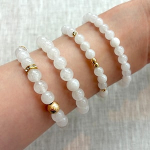 May include: Four white beaded bracelets with gold accents. One bracelet features a large gold bead, another has a gold-colored band with clear stones, and the other two have small gold beads. The bracelets are on a light background.