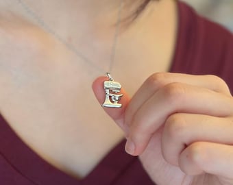 Puede incluir: Un collar con colgante plateado que representa una mini batidora de cocina. El dije detalla los componentes de la batidora, incluido el cuenco y el batidor. El collar se muestra sobre un fondo granate.