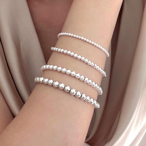 May include: Four silver beaded bracelets of varying sizes are stacked on an arm. The bracelets are made of small, round, shiny beads. The arm is positioned against a soft, neutral-colored background, creating a simple and elegant aesthetic.