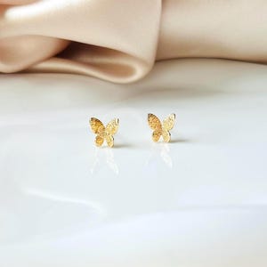 May include: A pair of small, gold-colored butterfly stud earrings. The earrings are set against a white surface with a soft, beige fabric in the background. The butterfly design has a textured surface, reflecting light.