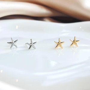 May include: Two pairs of starfish stud earrings. One pair is silver, the other gold. The earrings are presented on a white, reflective surface.
