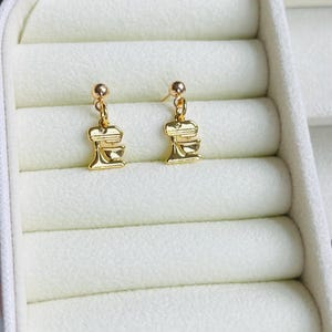 Könnte beinhalten: Ein Paar goldfarbene Ohrringe. Jeder Ohrring hat einen kleinen Anhänger in Form eines Küchenmixers, der an einem kleinen Kugelstecker befestigt ist. Die Ohrringe werden in einer weißen Schmuckschatulle mit weichem, strukturiertem Innenraum präsentiert.