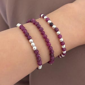 May include: Three beaded bracelets featuring faceted deep magenta gemstone beads alternating with smooth, polished silver-toned metallic beads. The bracelets are strung on elastic cord and worn together, showcasing a mix of textures and colors.