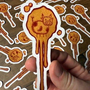 May include: A collection of stickers featuring a cartoon cow design. The stickers have a white border and a dripping, orange-yellow background. The cow is brown and orange. The stickers are held in a hand.