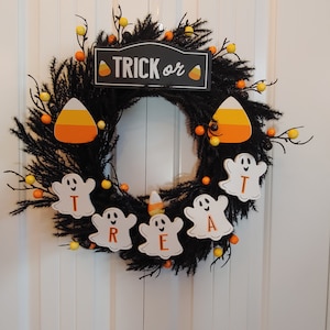 May include: A black Halloween wreath with a "Trick or Treat" sign and ghost-shaped ornaments with the letters "T", "R", "E", "A", "T" in orange. The wreath is decorated with yellow and orange candy corn and black berries.