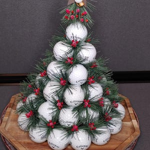 May include: A decorative Christmas tree made of golf balls, faux pine needles, and red berries. The tree is topped with a star and a golf club and ball. The tree sits on a wooden tray.