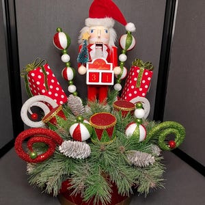 May include: A Christmas centerpiece featuring a nutcracker figurine holding a countdown calendar that says "Day 'til Christmas 31". The centerpiece is decorated with red and green ornaments, pine branches, and a red and gold drum.