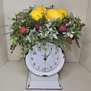 May include: A decorative vintage-style scale with a white face and black numbers, topped with a vibrant arrangement of yellow lemons, red strawberries, and green foliage. The scale has a distressed, rustic appearance.