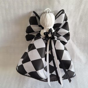May include: A decorative angel ornament featuring a white yarn head with a silver beaded halo. The body and wings are crafted from black and white checkered fabric. A small black fabric flower with a central rhinestone and thin ribbons adorn the front.