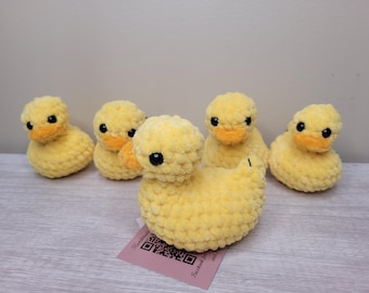 May include: Five handmade yellow crochet ducklings with orange beaks and black eyes. The ducklings are made of a soft, textured yarn. One duckling is larger than the others and is in the front. A pink tag with text and a QR code is in the front.