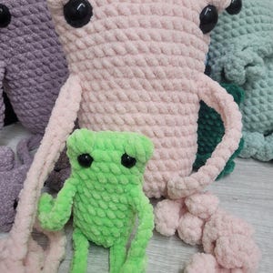 Könnte beinhalten: Zwei gehäkelte Frosch-Plüschtiere, eines rosa und eines grün. Der rosa Frosch hat große schwarze Augen und lange Arme. Der grüne Frosch ist kleiner und hat ein karikaturhaftes Aussehen.