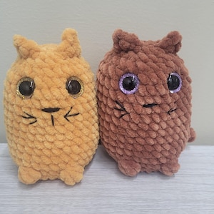 Op de afbeelding: Twee gehaakte pluche speelgoedjes in de vorm van katten. De kat links is geel en de kat rechts is bruin. Beide katten hebben grote zwarte ogen met glitter en een gestikte glimlach.