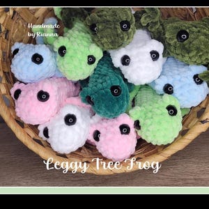 May include: A basket filled with handmade crocheted frog plushies in various colors, including green, blue, pink, and white. The frogs have black eyes and are sitting in a wicker basket. The text "Handmade by Rianna" and "Leggy Tree Frog" are visible on the image.