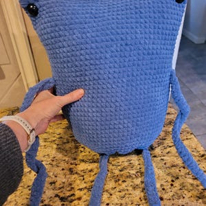 Puede incluir: Una gran criatura azul hecha a mano con ganchillo, con ojos de botón y largas extremidades colgantes. El peluche tiene una superficie texturizada y un diseño caprichoso, perfecto para una habitación infantil o como objeto decorativo único.