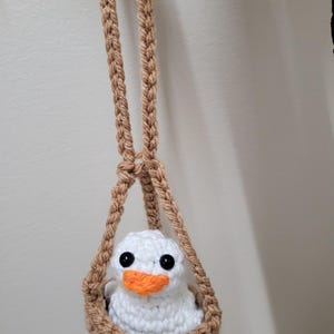 Peut inclure: Un caneton crocheté fait main dans un panier suspendu. Le caneton est blanc avec des yeux noirs et un bec orange. Le panier et les sangles suspendues sont de couleur marron clair. L'artisanat est suspendu sur un fond blanc.
