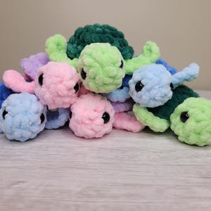 Könnte beinhalten: Eine Ansammlung handgefertigter, plüschiger Schildkrötenspielzeuge in verschiedenen Pastellfarben, darunter Rosa, Blau, Grün und Lila. Jede Schildkröte hat einen runden Kopf mit schwarzen Augen und einen strukturierten, flauschigen Körper. Die Schildkröten sind auf einer hellen Oberfläche gestapelt.
