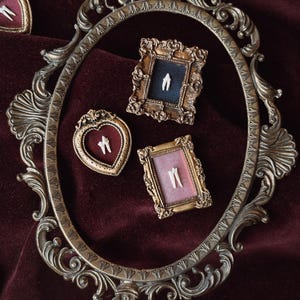 May include: A collection of small, ornate gold-toned frames containing white tooth specimens. One large oval frame surrounds a heart-shaped frame with a tooth, a rectangular frame with a dark blue background, and a rectangular frame with a pink background.