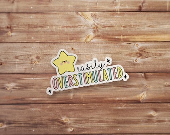 May include: A sticker with a yellow star character and the text "easily OVERSTIMULATED" in a colorful font. The sticker is on a wooden background.
