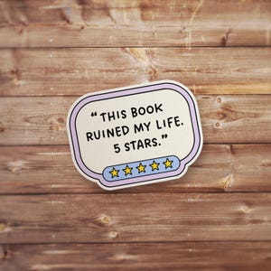 May include: A sticker with the text "THIS BOOK RUINED MY LIFE. 5 STARS." in black font. The sticker has a light purple border and a blue and yellow star rating at the bottom. The background is a wooden surface.
