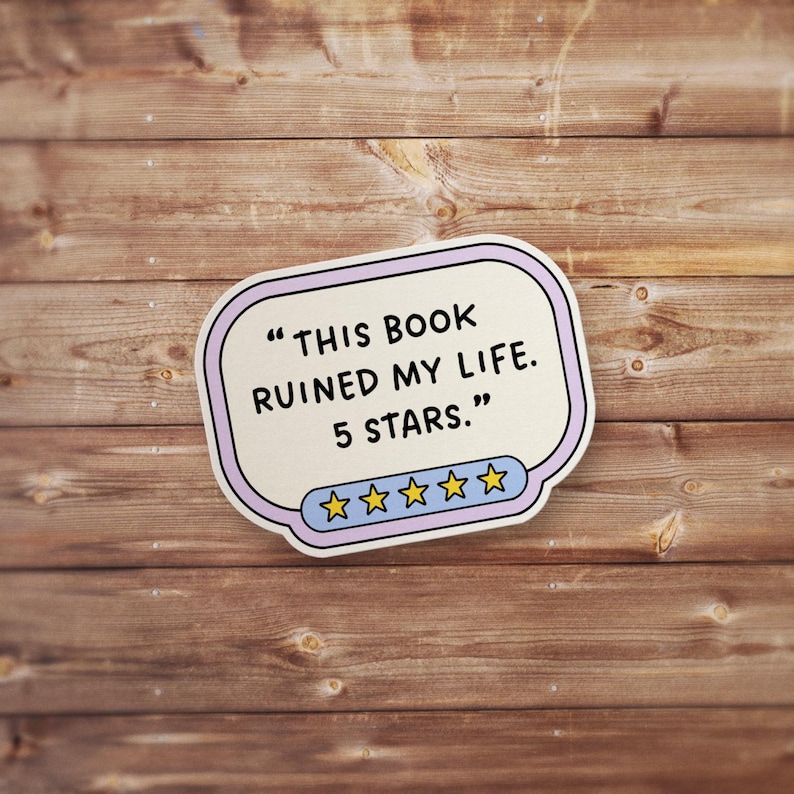 May include: A sticker with the text "THIS BOOK RUINED MY LIFE. 5 STARS." in black font. The sticker has a light purple border and a blue and yellow star rating at the bottom. The background is a wooden surface.