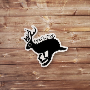 May include: A black sticker featuring a jackalope with antlers, running to the right. The text "STAY WEIRD" is above the jackalope. The sticker is on a wooden background.