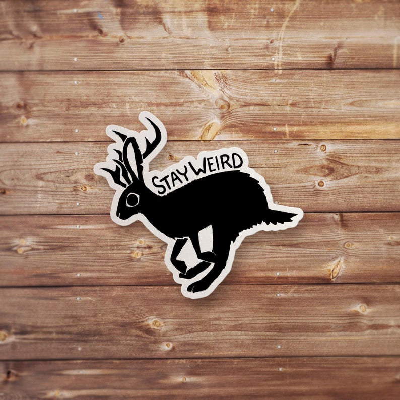 Peut inclure: Un autocollant noir repr&eacute;sentant un jackalope avec des bois, courant vers la droite. Le texte "STAY WEIRD" est au-dessus du jackalope. L'autocollant est sur un fond en bois.